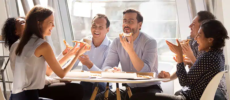 A group of employees sitting around a table laughing and eating pizza, engaging in a social wellbeing activity as part of a wider employee wellbeing strategy