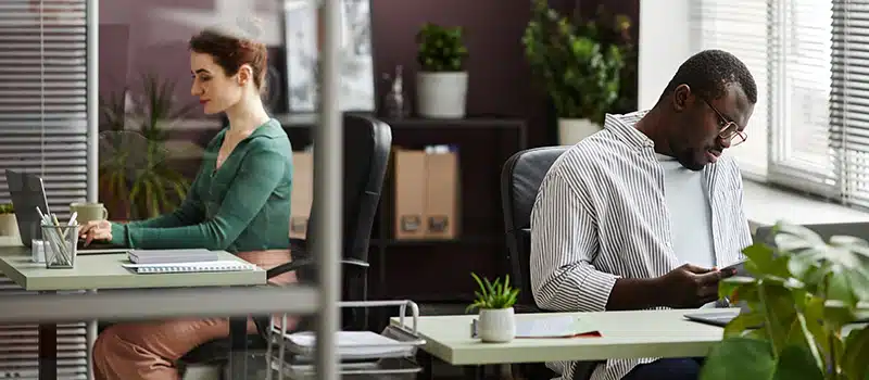 Two employees sitting at separate desks in a clean office with green plants dotted around, showcasing a good work environment as part of an employee wellbeing strategy