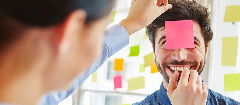 A woman sticking a sticky note to a smiling man's forehead as part of movember activities at work
