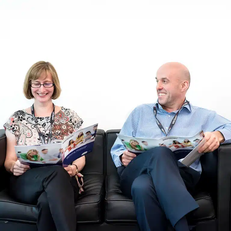 A man and a woman sitting on a black leather sofa smiling and reading a booklet as part of the New Leaf Health Employee Wellbeing Survey