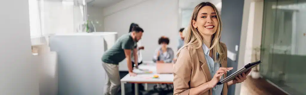 A woman in the workplace smiles whilst holding a tablet, with her colleagues out of focus in the background. Illustrative of understanding neurodiversity in the workplace
