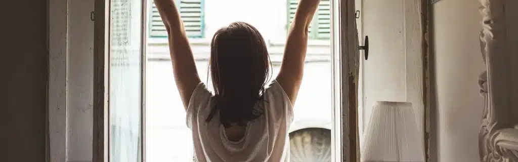 A woman stretching in front of an open window. Illustrative of how to become a morning person.