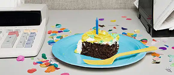 A piece of chocolate cake sitting on a colourful plate surrounded by confetti, on an office desk, next to an accountant's calculator.