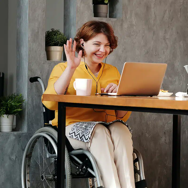 A woman in a wheelchair sitting at a desk waving at her laptop, attending a workplace wellbeing webinar.