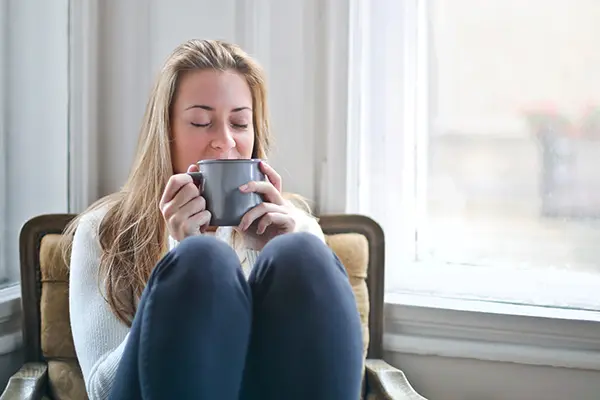 A woman sitting in a comfy chair, smiling with her eyes closed, drinking a hot drink from a mug, practising mindfulness techniques