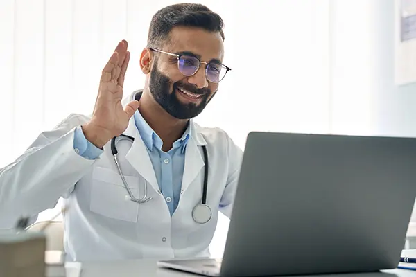 An online workplace GP sits at a desk and waves at a laptop screen, greeting a patient