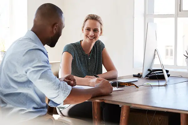 A man and a woman sitting at a desk talking and smiling