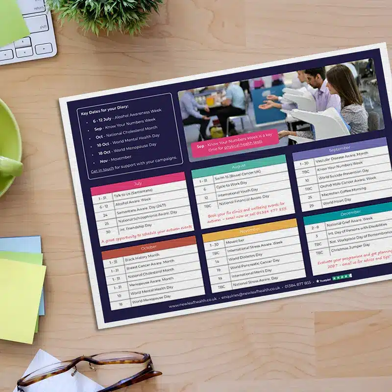 The 2026 Workplace Wellbeing Calendar from New Leaf Health on a desk next to some glasses, a keyboard, a mug, and a plant.