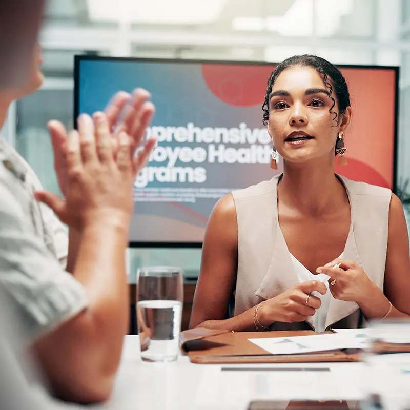 A woman sat in front of a TV delivering a presentation to colleagues on employee health and wellbeing
