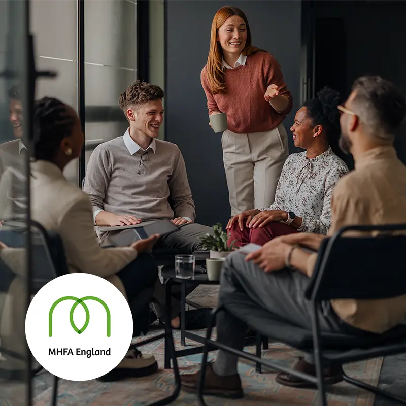A group of colleagues talking and smiling in a workplace, with the MHFA England logo in the bottom left corner