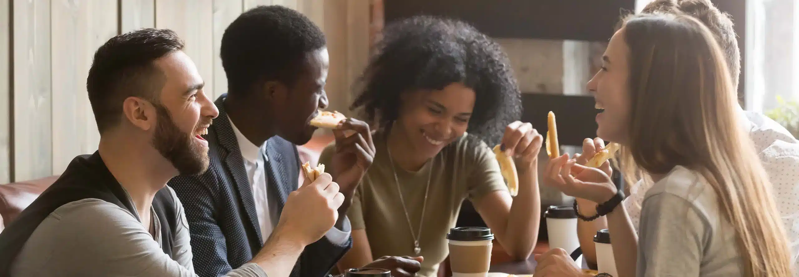 A group of people sitting at a table talking, smiling and eating pizza