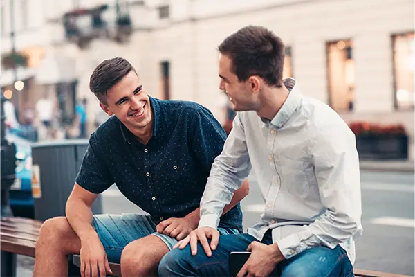 Two men sitting down and smiling at one another on a bench on a city street