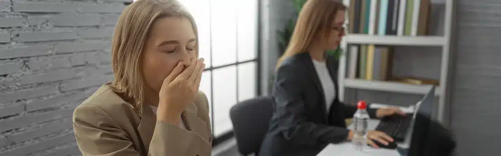 A woman in an office yawning sitting next to a woman on a laptop as an example of absenteeism and presenteeism