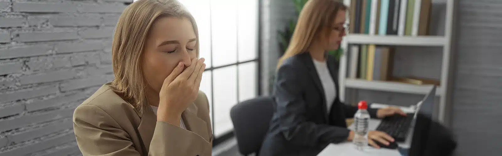 A woman in an office yawning sitting next to a woman on a laptop as an example of absenteeism and presenteeism