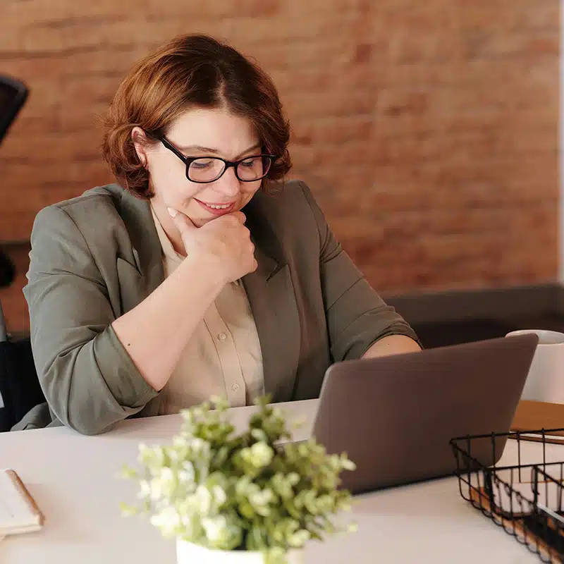 A woman with glasses sat smiling at a laptop on a desk