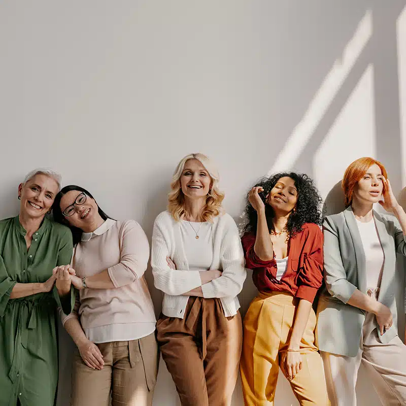 A group of diverse women standing against a white wall smiling at the camera