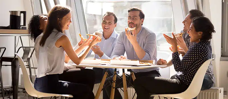 A group of employees engaging in a positive workplace wellbeing culture, eating pizza and laughing