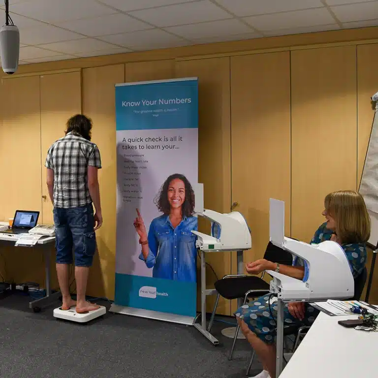 Two employees taking part in a New Leaf Health employee health check event, using body composition scales and a blood pressure machine