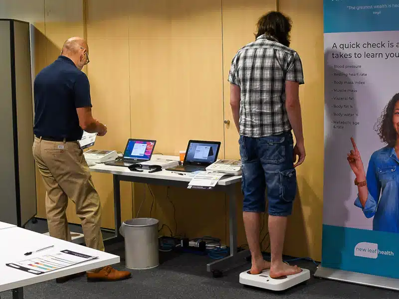 A man standing on a set of body composition scales as part of an employee health check event, with a New Leaf Health practitioner standing next to him