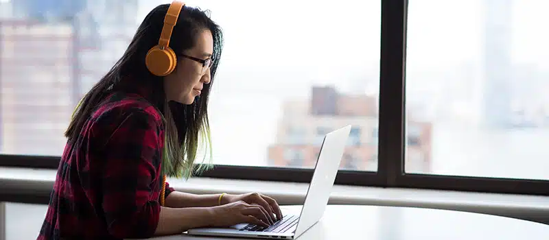 A woman wearing headphone sat in front of an office window on a laptop completing an employee wellbeing survey
