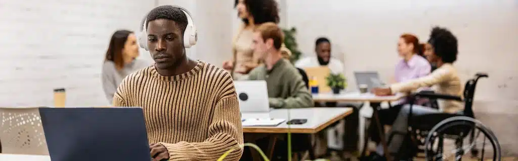 A man sits working on a laptop in the foreground weaing headphones, while a group of diverse colleagues work in the background, showcasing neurodiversity at work.