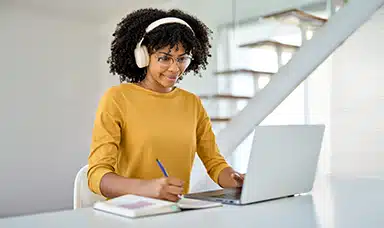 A woman wearing headphones working on a laptop and writing in a journal, representing neurodiversity at work