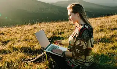 A woman sat on a hill working on her laptop in the sunshine