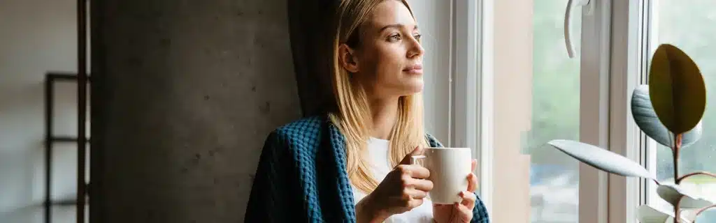 A woman experiencing seasonal affective disorder symptoms wrapped in a blanket staring out of a window drinking a hot drink from a mug